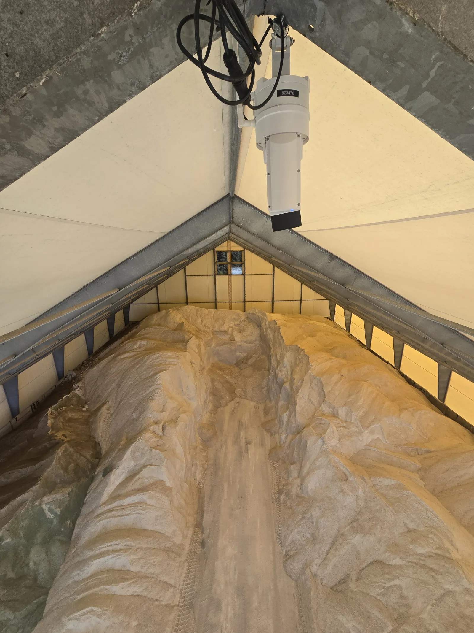 EOS2 LiDAR sensor mounted at the ridge of a fabric-roofed salt barn, looking down the length of a road salt pile pushed up against both side walls with a loader lane down the middle