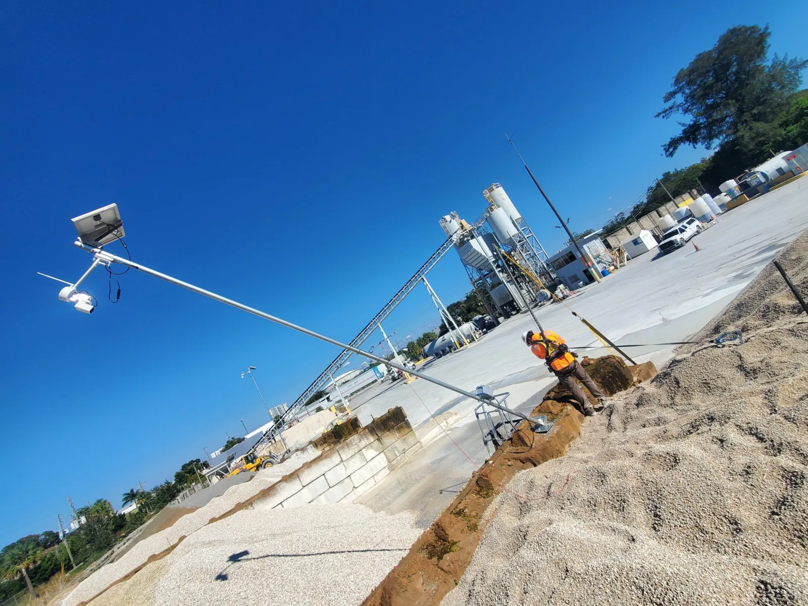 Technician raising a solar-powered EOS2 LiDAR sensor pole on a hinged mounting base above aggregate stockpiles at a ready-mix plant