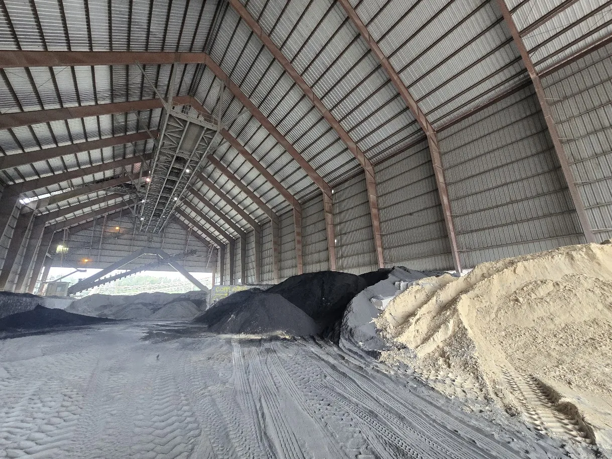 Coal, additive and raw material stockpiles side by side in an enclosed cement plant storage building, with a conveyor gallery overhead