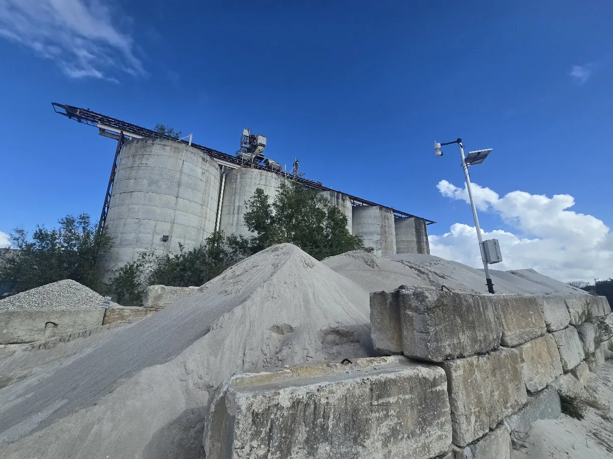 Row of concrete silos behind sand and aggregate stockpiles, with a solar-powered EOS2 LiDAR sensor on a pole