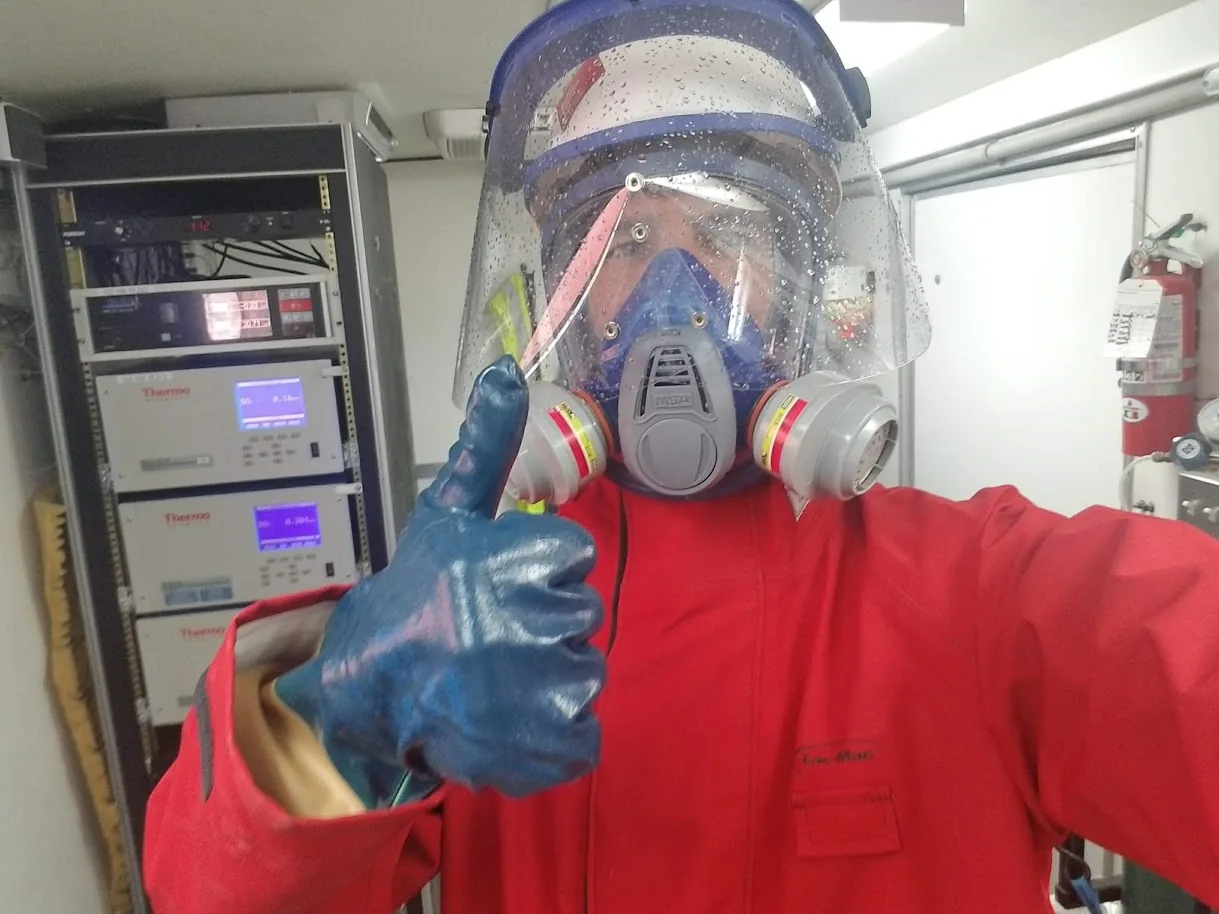 Technician in a full-face respirator, face shield and acid-resistant suit giving a thumbs up beside a rack of gas analyzers in a CEMS shelter